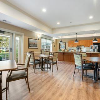 A spacious dining area with wooden flooring, featuring multiple tables and chairs, adjacent to a modern kitchen.
