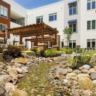 A modern apartment complex features a serene outdoor garden with a pergola and a small stream flowing through rocks.
