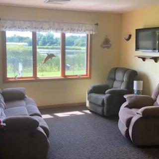 A cozy living room with large windows offering a scenic lake view, featuring comfortable seating and a wall-mounted television.