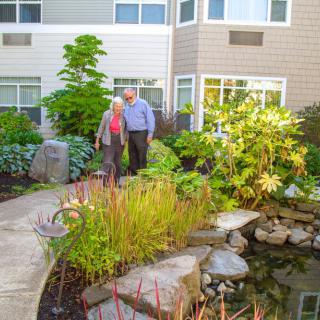 An elderly couple enjoys a walk through a lush garden with a pond in a residential area.