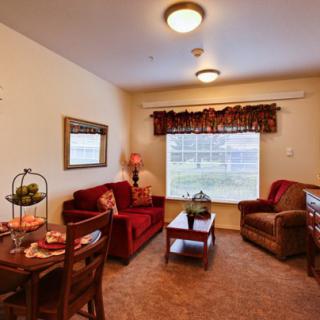 A cozy living room with a sofa, armchair, and dining area, featuring warm tones and a large window overlooking greenery.