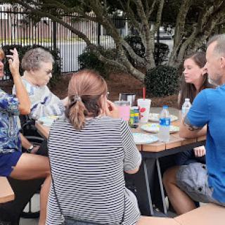 A group of friends enjoying a lively conversation over food and drinks at an outdoor table.