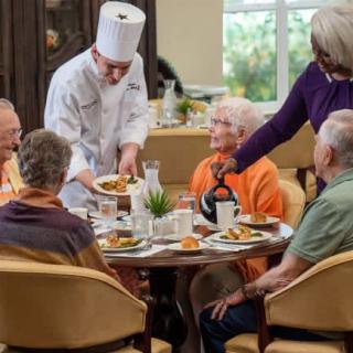 A chef serves meals to a group of seniors dining together in a communal setting.