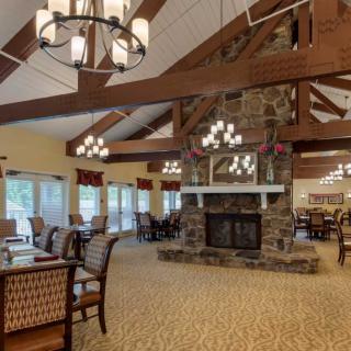 A spacious dining and lounge area with a central fireplace, high ceilings, chandeliers, and wooden beams.