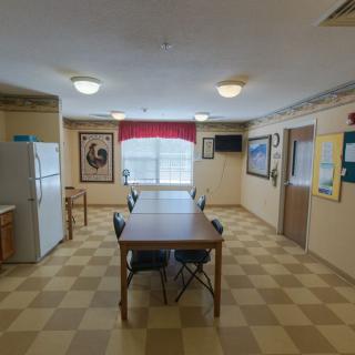A communal dining area with a checkered floor, featuring a table and chairs, refrigerator, window with curtains, and various artworks on the walls.