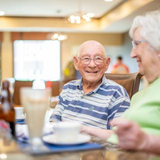 Two senior individuals enjoying a conversation over a meal in a bright dining area.