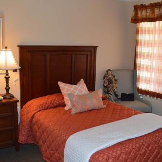 A cozy bedroom with a red quilted bedspread, wooden headboard, bedside table with a lamp, and a chair by the window.