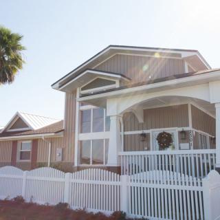 A two-story house with a porch and white fence, illuminated by sunlight.