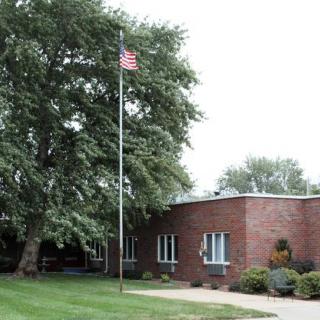 A brick building with an American flag on a flagpole, surrounded by trees and grass.