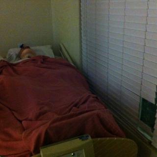 A hospital room with a patient lying in bed covered by a blanket, next to a window with blinds.