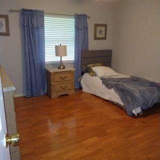 A simple bedroom with hardwood floors, featuring a single bed, nightstand, and blue curtains.