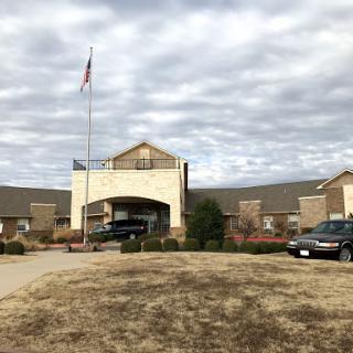 A welcoming entrance of a building under a cloudy sky, featuring a flag and vehicles in the parking area.