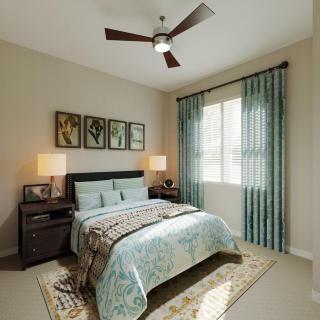 A cozy bedroom interior featuring a neatly made bed with patterned bedding, two bedside lamps, wall artwork, light curtains, and a ceiling fan.