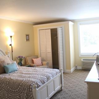 A cozy and well-lit bedroom featuring a bed, floor lamp, and a wardrobe with an open closet door. The room has light-colored walls and carpeted flooring.