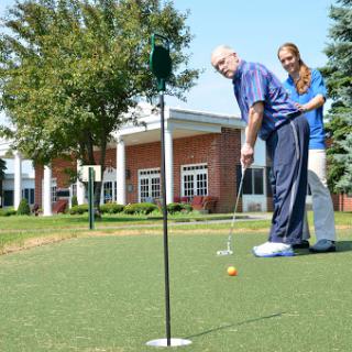 A young person assists an elderly individual with putting on a golf green.