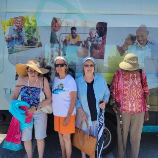 A group of seniors posing together in front of a vehicle, enjoying a sunny day out.