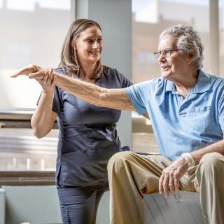 A therapist assists an elderly man with arm exercises during a physical therapy session.