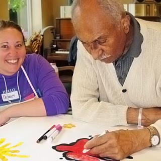 A joyful art session where an elderly man is engaged in painting, assisted by a smiling caregiver.