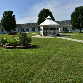 A neatly maintained lawn with a central gazebo, surrounded by trees and a building in the background.