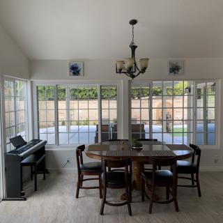A bright dining room with a table, chairs, and a piano, illuminated by natural light through large windows.