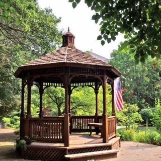 A wooden gazebo in a lush garden setting, surrounded by greenery.