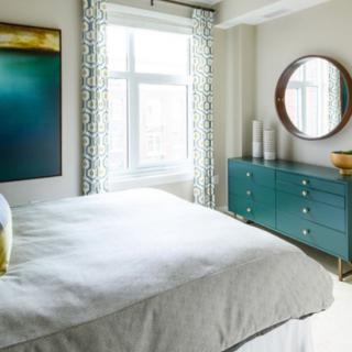 A modern bedroom featuring a comfortable bed, large window, abstract artwork, and a stylish dresser with a round mirror.