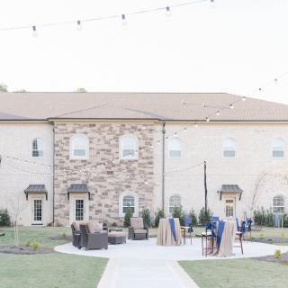 A building with a stone facade features outdoor seating on a patio, adorned with string lights, and surrounded by a well-maintained lawn.