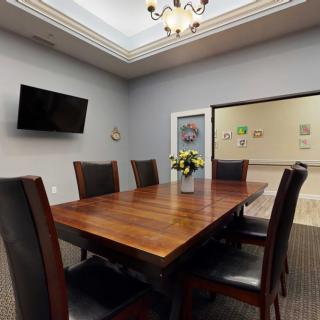 A modern conference room with a wooden table, six chairs, a wall-mounted television, and floral centerpiece.