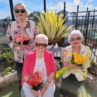 Three women wearing sunglasses hold colorful flowers while sitting in a sunny garden.