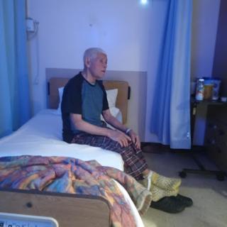 An elderly man sitting on the edge of a bed in a hospital room, illuminated by soft blue light.