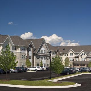 A modern residential apartment complex with a parking lot and trees under a clear blue sky.