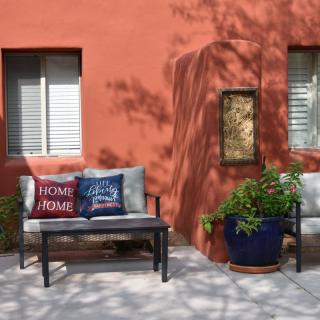 A cozy patio area with a bench, decorative cushions, and potted plants against a warm-colored wall.