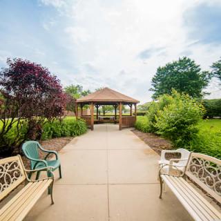 A peaceful garden with a pathway leading to a wooden gazebo, surrounded by lush greenery and benches.