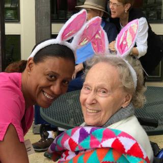 Two individuals wearing bunny ears are smiling outdoors, one wrapped in a colorful blanket.