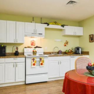 A cozy kitchen with white cabinets, a stove, and a dining area featuring a fruit bowl on a red tablecloth.