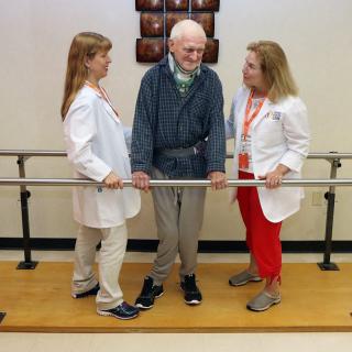 A senior man receives assistance from two healthcare professionals during a physical therapy session.