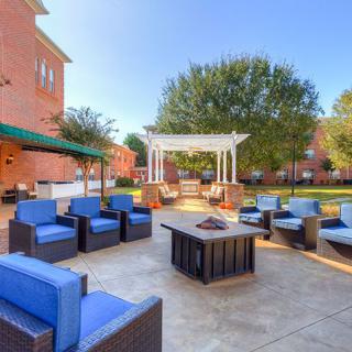 A spacious outdoor patio area featuring comfortable seating with blue cushions, a central fire pit, and a gazebo set against a backdrop of trees and brick buildings.