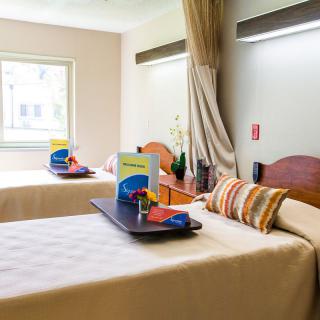 A hospital room featuring two patient beds, each with a tray of items by the window.