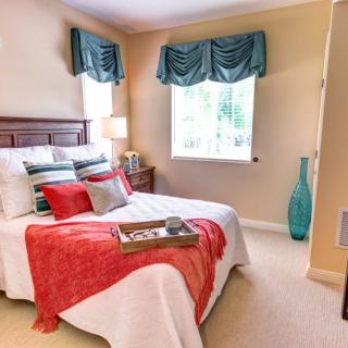 A cozy bedroom featuring a neatly made bed with colorful pillows and a red blanket, complemented by a stylish lamp and a tall blue vase near the window.