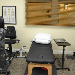 A therapy room equipped with a treatment table, exercise machine, and anatomical poster.