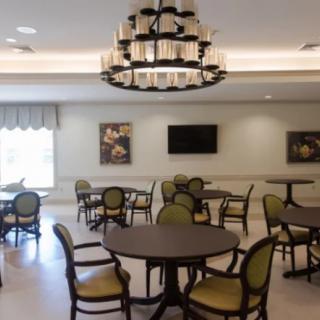 A spacious dining room featuring round tables and cushioned chairs, illuminated by a large chandelier.