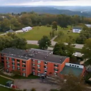 An aerial view of a red brick building surrounded by greenery and trees under an overcast sky.
