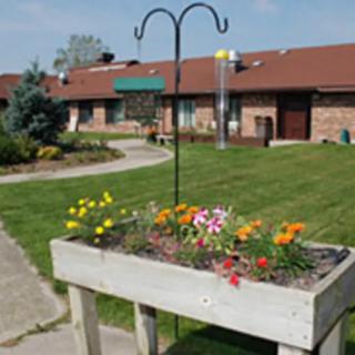 A raised flower bed with colorful flowers in front of a brick building and well-maintained lawn.
