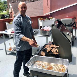 A chef grilling meat on a barbecue in an outdoor setting.