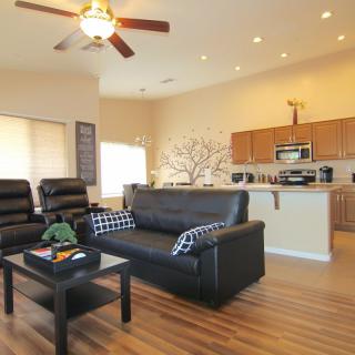 A modern open-plan living room and kitchen with stylish furnishings, featuring a black couch, wood flooring, and a ceiling fan.