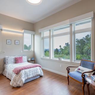 A cozy bedroom with a large window, allowing ample natural light. The room features a single bed, wooden flooring, and an elegant chair.
