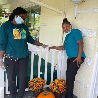 Two individuals making a delivery on a porch decorated with flowers and a pumpkin, both wearing masks and gloves.