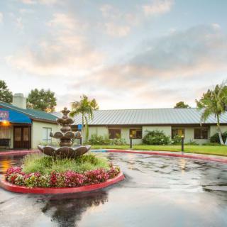 A scenic view of a building with a circular driveway, featuring a central fountain surrounded by colorful flowers and palm trees at sunset.