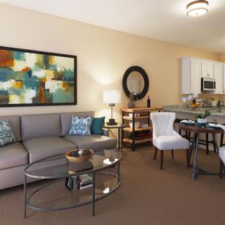 A modern living room featuring a comfortable sofa, glass coffee table, abstract artwork, and a small dining area connected to the kitchen.