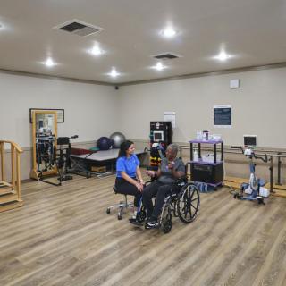 A therapist assists a patient in a wheelchair within a rehabilitation facility equipped with various exercise machines and therapy tools.